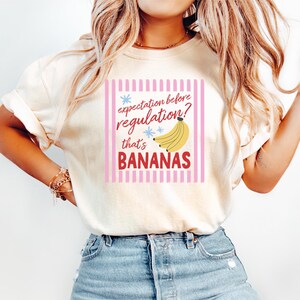 Peut inclure: Un t-shirt à manches courtes de couleur crème avec un imprimé graphique à rayures verticales roses. Le texte "expectation before regulation? that's BANANAS" est écrit en rouge et noir, avec une illustration jaune d'une grappe de bananes.