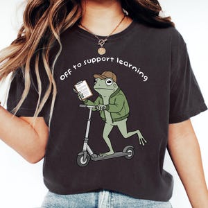 Puede incluir: Camiseta gris oscuro con un dibujo animado de una rana en un patinete, sosteniendo una carpeta. La rana lleva una chaqueta verde y una gorra marrón. El texto encima de la rana dice "OFF to support learning."