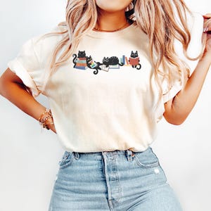 Puede incluir: Una camiseta de manga corta de color crema con un estampado gráfico de cuatro gatos negros interactuando con pilas de libros coloridos. Los gatos aparecen en varias posturas, incluyendo sentados sobre, tumbados encima y leyendo los libros. La camiseta se combina con unos vaqueros de mezclilla azul claro.
