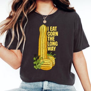 Op de afbeelding: Een antracietgrijs T-shirt met korte mouwen voorzien van een afbeelding van gele maïskolven en de tekst I EAT CORN THE LONG WAY in het geel. Het ontwerp bevat een hoge verticale maïskolf en twee kleinere horizontale maïsstukken met groene bladeren.