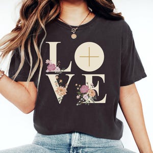 May include: Dark gray t-shirt with the word "LOVE" in large, cream-colored letters. The "O" is a circle with a cross inside. Floral arrangements in pink, purple, and yellow decorate the letters. The shirt is worn by a person wearing a gold necklace and bracelet.