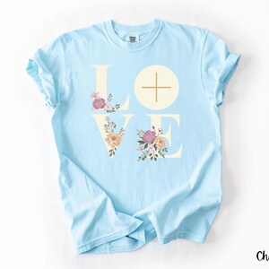 May include: Pale blue t-shirt featuring the word "LOVE" in cream, with floral designs within the letters. The "L" and "E" are adorned with pink, peach, and green flowers. A cross is in the centre of the "O". The word "Chambray" is in the bottom right corner.