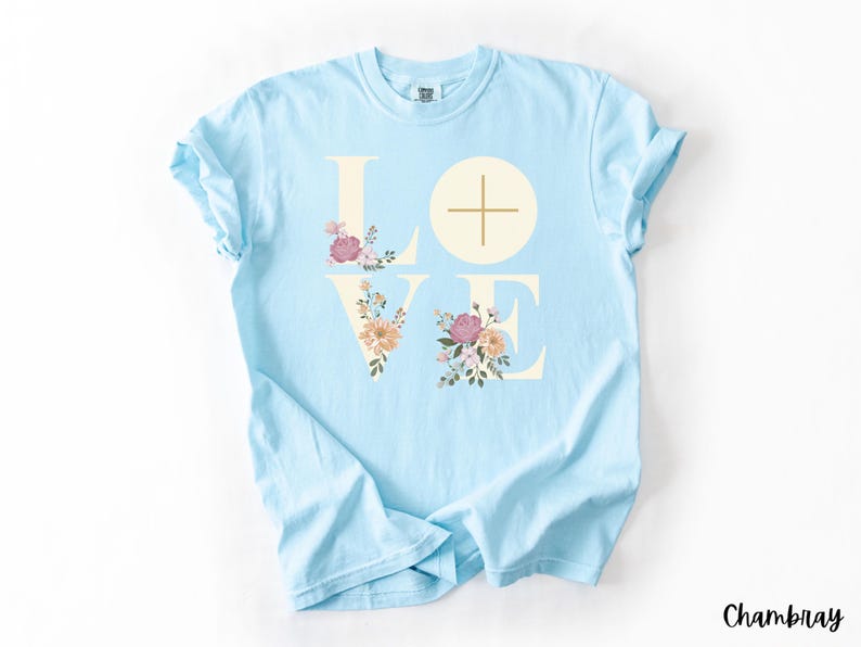 May include: Pale blue t-shirt featuring the word "LOVE" in cream, with floral designs within the letters. The "L" and "E" are adorned with pink, peach, and green flowers. A cross is in the centre of the "O". The word "Chambray" is in the bottom right corner.
