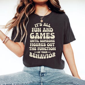 May include: Dark grey t-shirt featuring the text "IT'S ALL FUN AND GAMES UNTIL SOMEONE FIGURES OUT THE FUNCTION OF YOUR BEHAVIOR" in a retro font. The shirt is worn with blue jeans. A casual wear item.