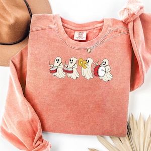 May include: A coral-coloured crewneck sweatshirt featuring a graphic of four white ghosts playing musical instruments, including drums and cymbals. The garment has a ribbed collar with a Comfort Colors label visible at the inner neckline.