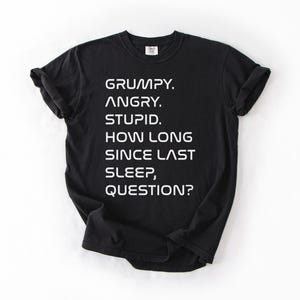 May include: Black t-shirt with white text. The text reads: "GRUMPY. ANGRY. STUPID. HOW LONG SINCE LAST SLEEP, QUESTION?" The shirt is a crew neck style.