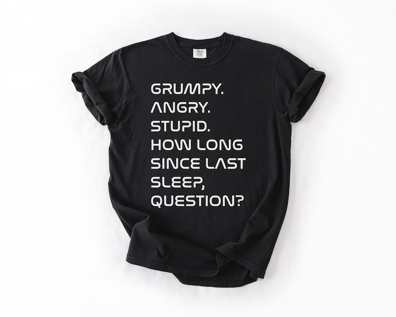 May include: Black t-shirt with white text. The text reads: "GRUMPY. ANGRY. STUPID. HOW LONG SINCE LAST SLEEP, QUESTION?" The shirt is a crew neck style.