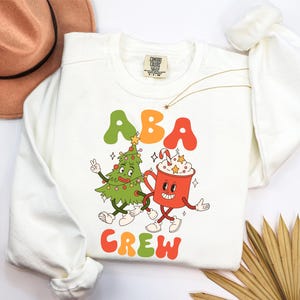 May include: A white long-sleeve sweatshirt featuring a graphic print of a cartoon Christmas tree and a red mug of hot cocoa with the text ABA CREW in colorful, bubbly letters. The design includes stars, a candy cane, and festive lights.