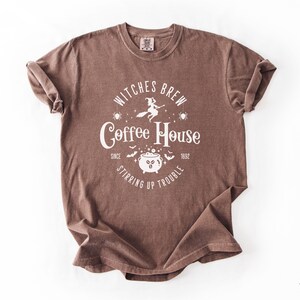 以下が含まれることがあります： 茶色の半袖Tシャツ。白のグラフィックプリントで「Witches Brew Coffee House Since 1692 Stirring Up Trouble」と書かれており、ほうきに乗った魔女、泡立つ大釜、クモ、コウモリのイラストが描かれています。