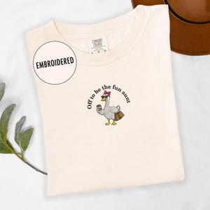 May include: A cream-coloured t-shirt featuring an embroidered design of a goose wearing sunglasses and a pink bow, holding a coffee cup and a bag. The text "Off to be the fun aunt" is embroidered around the goose. The shirt has a tag that says "Embroidered".