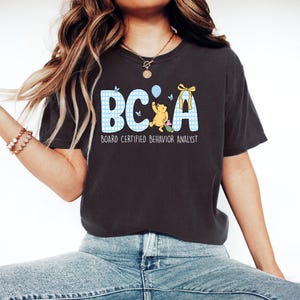 Op de afbeelding: Een antracietgrijs T-shirt met korte mouwen en een afbeelding met de letters BC en A in een blauw-wit ruitpatroon. Het ontwerp bevat een gele beer die een blauwe ballon vasthoudt, een klein roze figuurtje en de tekst BOARD CERTIFIED BEHAVIOR ANALYST.