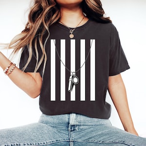 May include: Black t-shirt with a referee-style design. The shirt features vertical white stripes and a black and white whistle pendant. The design is centered on the front of the shirt, creating a bold, graphic look.