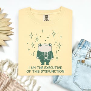 May include: A pale yellow t-shirt featuring a graphic of a cartoon frog wearing a dark green suit and tie. The frog is surrounded by small star icons. Below the frog, the text reads, "I AM THE EXECUTIVE OF THIS DYSFUNCTION." The shirt has a Comfort Colors label.