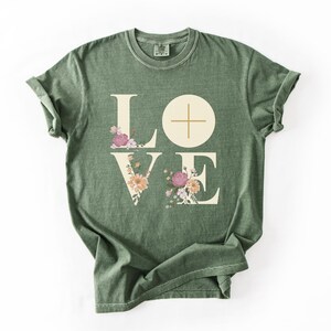 May include: Moss green t-shirt featuring the word "LOVE" in large, cream-coloured letters. The "L" and "E" are decorated with floral arrangements in pink, peach, and cream. The "O" is a cream-coloured circle with a cross.