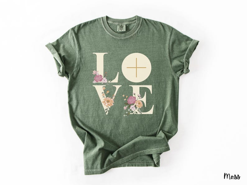 May include: Moss green t-shirt featuring the word "LOVE" in large, cream-coloured letters. The "L" and "E" are decorated with floral arrangements in pink, peach, and cream. The "O" is a cream-coloured circle with a cross.
