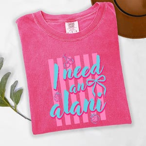 May include: A bright pink short-sleeve t-shirt with a crew neckline. The front features a graphic with vertical light pink stripes and the text I need an alani in light blue script. The design includes small line art of cherries and strawberries.