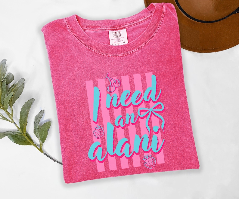 May include: A bright pink short-sleeve t-shirt with a crew neckline. The front features a graphic with vertical light pink stripes and the text I need an alani in light blue script. The design includes small line art of cherries and strawberries.