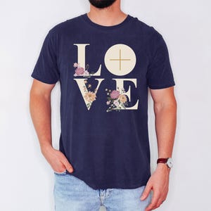 May include: A navy blue t-shirt featuring the word "LOVE" in a cream font. The letters "L" and "V" are decorated with floral designs in pink, peach, and cream. A cream circle with a cross is above the "E".