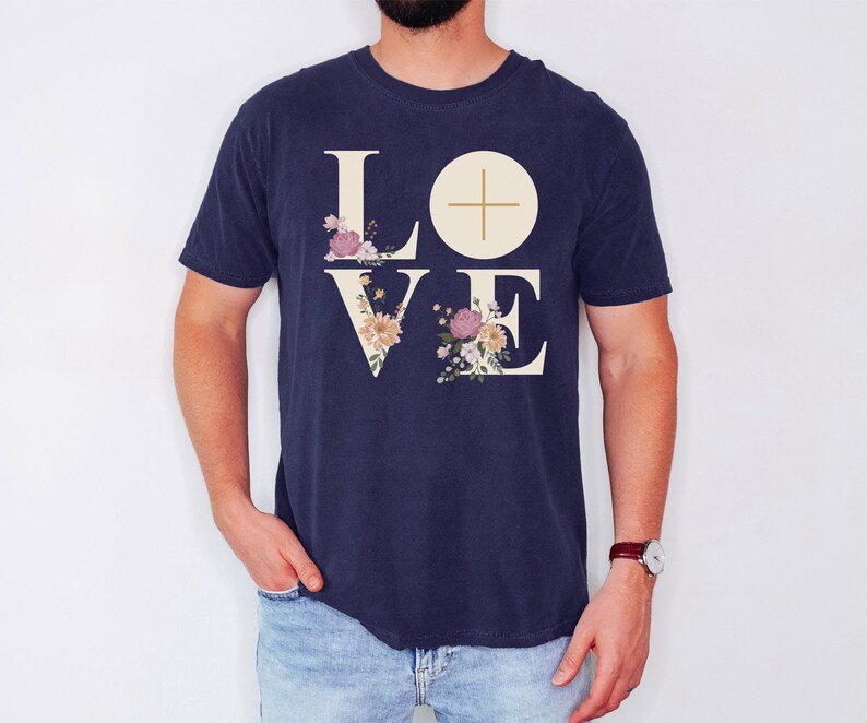 May include: A navy blue t-shirt featuring the word "LOVE" in a cream font. The letters "L" and "V" are decorated with floral designs in pink, peach, and cream. A cream circle with a cross is above the "E".
