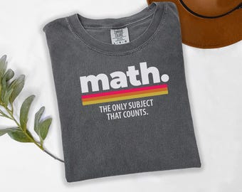 May include: A charcoal gray crew neck t-shirt featuring the white text math. above three horizontal stripes in pink, orange, and yellow. Below the stripes, the text reads THE ONLY SUBJECT THAT COUNTS. The shirt has a Comfort Colors brand label.