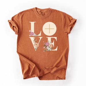 May include: A terracotta-coloured t-shirt featuring the word "LOVE" in large, beige block letters. The "L" and "E" are decorated with floral arrangements in pink, purple, and cream. The "O" is a circle with a cross.