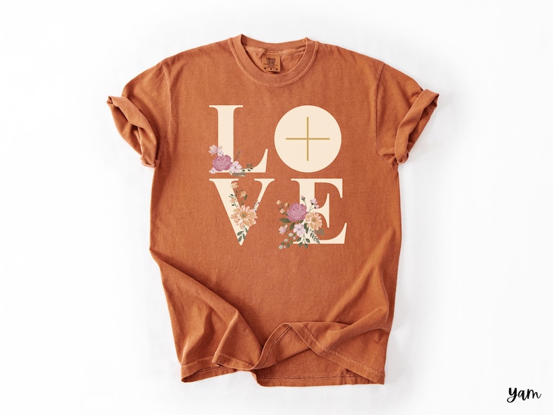 May include: A terracotta-coloured t-shirt featuring the word "LOVE" in large, beige block letters. The "L" and "E" are decorated with floral arrangements in pink, purple, and cream. The "O" is a circle with a cross.