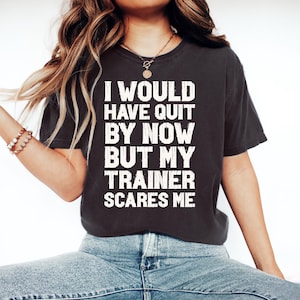 Pode incluir: Camiseta cinza escuro com o texto branco: "I WOULD HAVE QUIT BY NOW BUT MY TRAINER SCARES ME". A camiseta é combinada com jeans azuis e um colar de ouro.