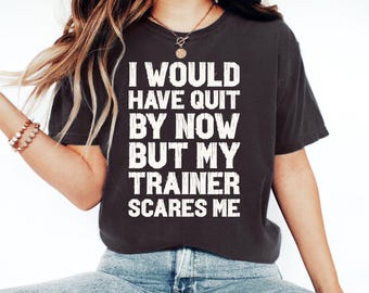 Peut inclure: T-shirt gris foncé avec l'inscription blanche : « I WOULD HAVE QUIT BY NOW BUT MY TRAINER SCARES ME ». Le t-shirt est porté avec un jean bleu et un collier doré.