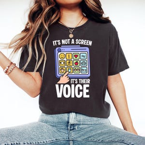 May include: Dark gray t-shirt with the text "IT'S NOT A SCREEN IT'S THEIR VOICE". The shirt features a graphic of a tablet with various icons, including a smiley face, heart, and car. A finger is pointing at the tablet.