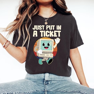 May include: Dark grey t-shirt featuring the text "JUST PUT IN A TICKET" above a retro cartoon computer character. The computer is smiling and waving, set against a sunset-coloured circle.