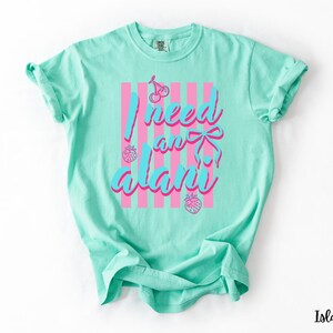 May include: A mint green short-sleeve t-shirt featuring a graphic print with vertical pink stripes, two cherries, two strawberries, and a blue bow. The text reads I need an alani in a blue script font with a pink outline centred on the front.
