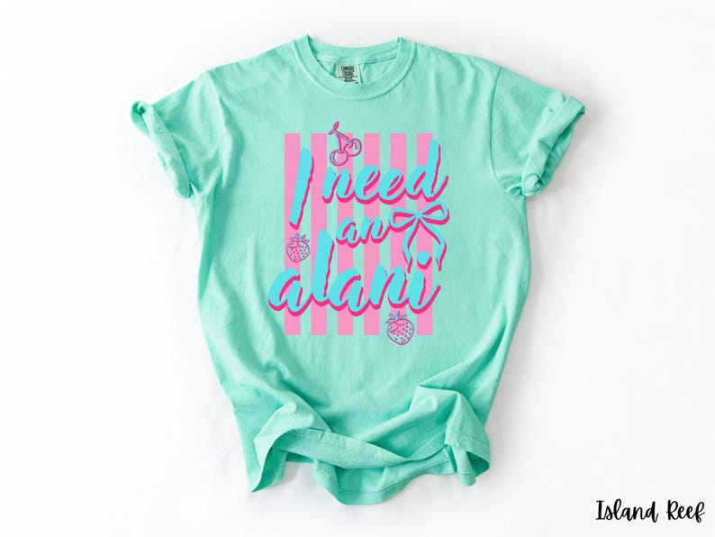 May include: A mint green short-sleeve t-shirt featuring a graphic print with vertical pink stripes, two cherries, two strawberries, and a blue bow. The text reads I need an alani in a blue script font with a pink outline centred on the front.