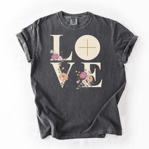 May include: Dark grey t-shirt featuring the word "LOVE" in large, off-white letters. The "O" is a circle with a cross. Floral arrangements in pink, peach, and white decorate the letters. The word "Pepper" is in the bottom right corner.