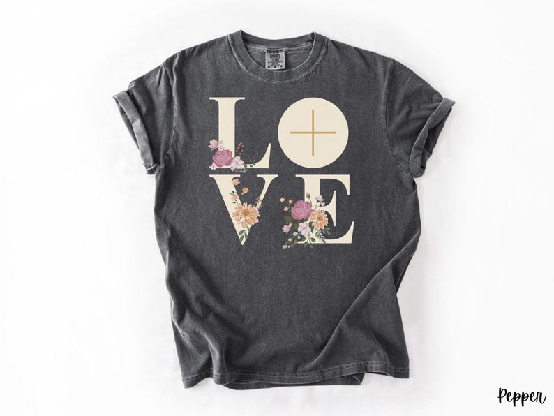 May include: Dark grey t-shirt featuring the word "LOVE" in large, off-white letters. The "O" is a circle with a cross. Floral arrangements in pink, peach, and white decorate the letters. The word "Pepper" is in the bottom right corner.