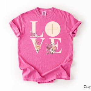 May include: A vibrant pink t-shirt featuring the word "LOVE" in a light beige font, adorned with floral designs. The letter "O" is a circle with a cross. The brand name "Crunchberry" is printed in the bottom right corner. The t-shirt is a comfortable, casual wear item.