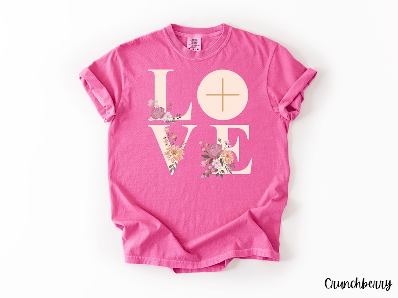 May include: A vibrant pink t-shirt featuring the word "LOVE" in a light beige font, adorned with floral designs. The letter "O" is a circle with a cross. The brand name "Crunchberry" is printed in the bottom right corner. The t-shirt is a comfortable, casual wear item.