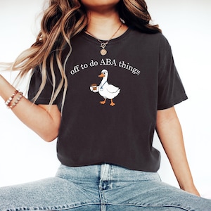 May include: A charcoal gray t-shirt featuring a white graphic of a goose wearing glasses and a blue lanyard while holding a coffee cup. The text above the goose reads, off to do ABA things, in a white serif font. The shirt has short sleeves.