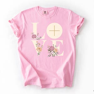 May include: A pale pink t-shirt featuring the word "LOVE" in a cream hue. The "O" is a circle with a cross. Floral designs in pink, peach, and green adorn the letters. The word "Blossom" is written in black in the bottom right corner.