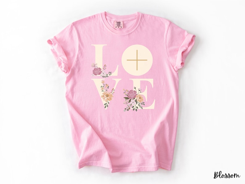 May include: A pale pink t-shirt featuring the word "LOVE" in a cream hue. The "O" is a circle with a cross. Floral designs in pink, peach, and green adorn the letters. The word "Blossom" is written in black in the bottom right corner.