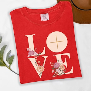 May include: A red t-shirt featuring the word "LOVE" in cream, decorated with floral designs. The letter "O" is a circle with a cross. The shirt is folded on a white surface.
