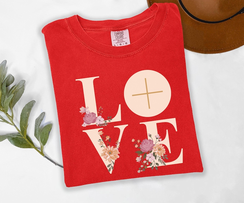 May include: A red t-shirt featuring the word "LOVE" in cream, decorated with floral designs. The letter "O" is a circle with a cross. The shirt is folded on a white surface.