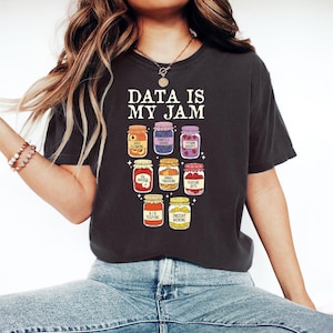 Può includere: T-shirt grigio scuro con la scritta "DATA IS MY JAM" sopra una grafica di nove barattoli di marmellata, ciascuno etichettato con termini di data science come "DATA CLEANING" e "PYTHON SCRIPTS". La maglietta è realizzata in materiale morbido.