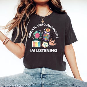 Op de afbeelding: Donkergrijs t-shirt met de tekst "HOWEVER YOU COMMUNICATE I'M LISTENING". Het shirt heeft kleurrijke afbeeldingen van een hoortoestel, een rekenmachine, een hart en een gebarentaal hand symbool. Het shirt is gemaakt van een zacht materiaal.