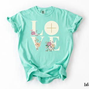 May include: A turquoise t-shirt featuring the word "LOVE" in off-white capital letters. The "O" is a circle with a cross. Floral designs in pink, peach, and green decorate the letters. The shirt is a relaxed, casual fit.