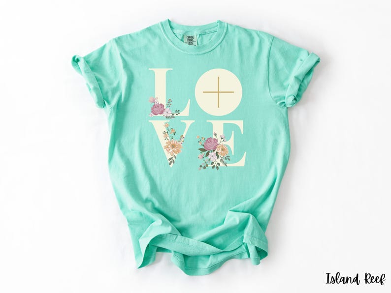 May include: A turquoise t-shirt featuring the word "LOVE" in off-white capital letters. The "O" is a circle with a cross. Floral designs in pink, peach, and green decorate the letters. The shirt is a relaxed, casual fit.