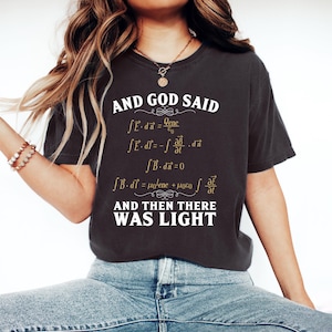 May include: A charcoal gray short-sleeve t-shirt featuring white and gold text that reads AND GOD SAID followed by Maxwell's equations and the text AND THEN THERE WAS LIGHT. The shirt has a crew neckline and a relaxed fit with a vintage-washed appearance.