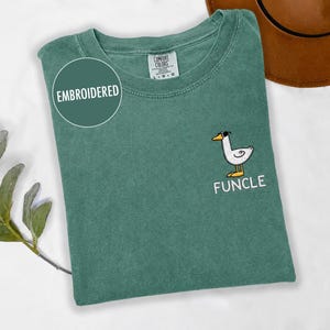 May include: A teal green t-shirt with an embroidered design of a white duck wearing sunglasses and the word "FUNCLE" below. The shirt has a crew neck and short sleeves. The word "EMBROIDERED" is in a circle on the shirt.