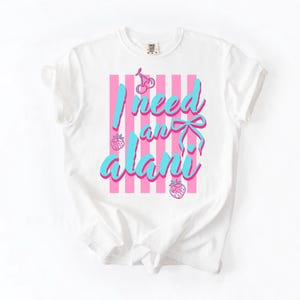 May include: A white short-sleeved t-shirt featuring a graphic design with vertical pink stripes. The text "I need an alani" is printed in a bright blue script font, accompanied by illustrations of cherries, a bow, and two strawberries.