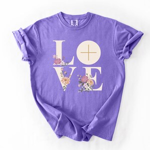 May include: A violet t-shirt featuring the word "LOVE" in a white, stylized font. The "O" is a circle with a cross, and the letters are decorated with floral arrangements. The word "Violet" is printed at the bottom right.