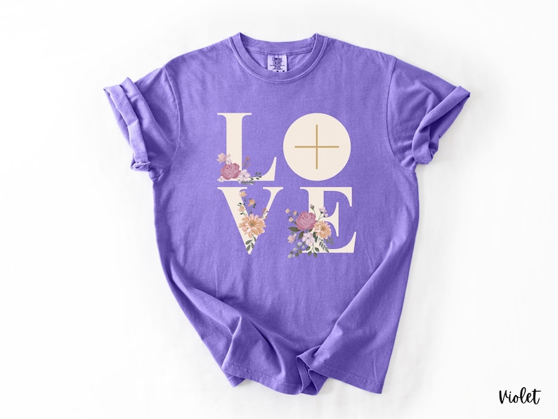 May include: A violet t-shirt featuring the word "LOVE" in a white, stylized font. The "O" is a circle with a cross, and the letters are decorated with floral arrangements. The word "Violet" is printed at the bottom right.